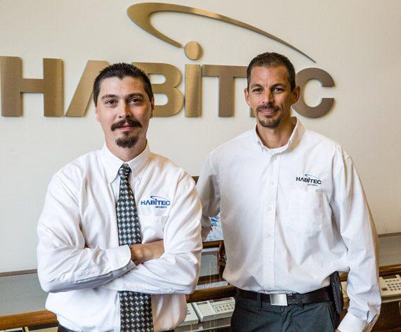 Two members of the Habitec Security team in front of the logo of the company mounted on a wall